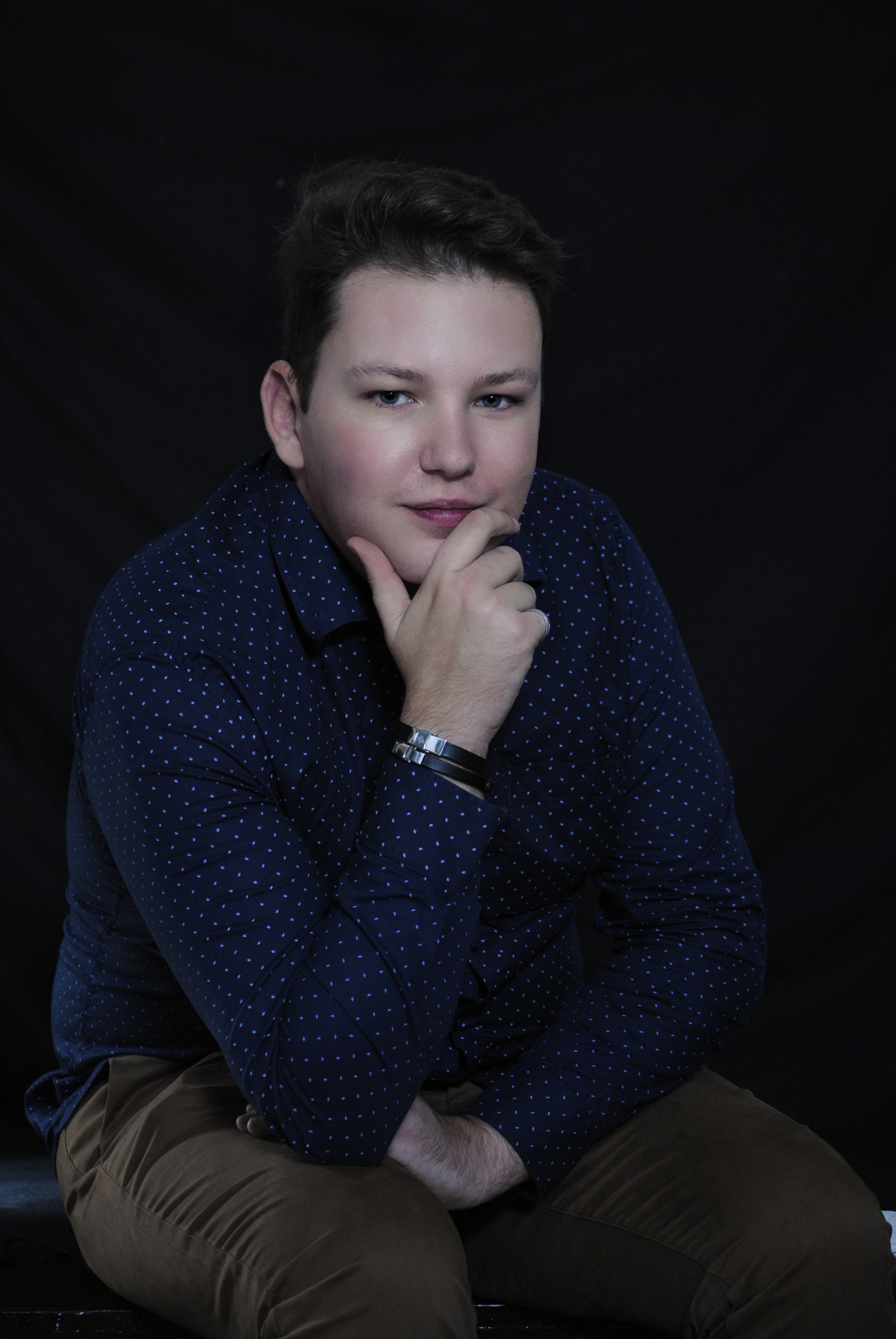 Portrait of a person sitting with their hand resting on their chin, looking thoughtfully into the camera. They are wearing a navy blue shirt with small white polka dots and brown pants. The background is dark, highlighting the subject's face and upper body.